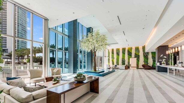 A modern hotel lobby with large windows, a central seating area, a reception desk, and a green wall with vertical gardens.
