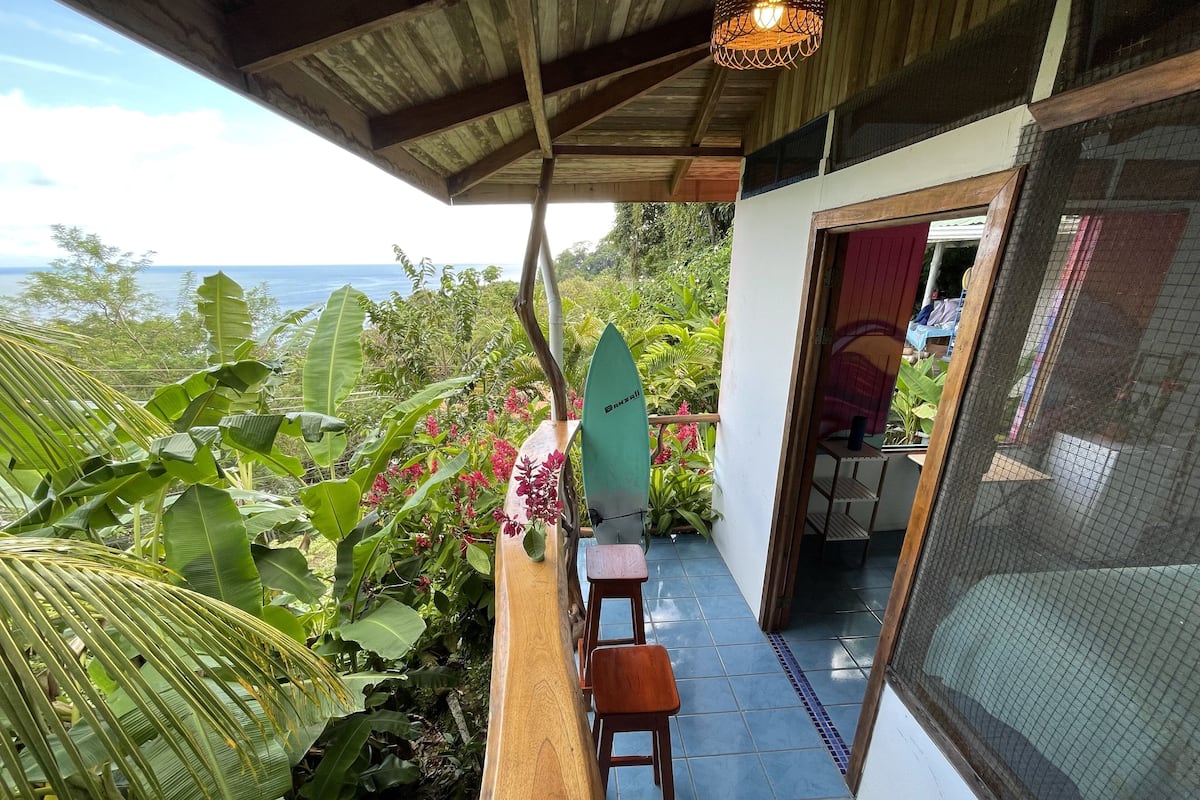 A balcony with a wooden bench, a surfboard, and lush greenery.