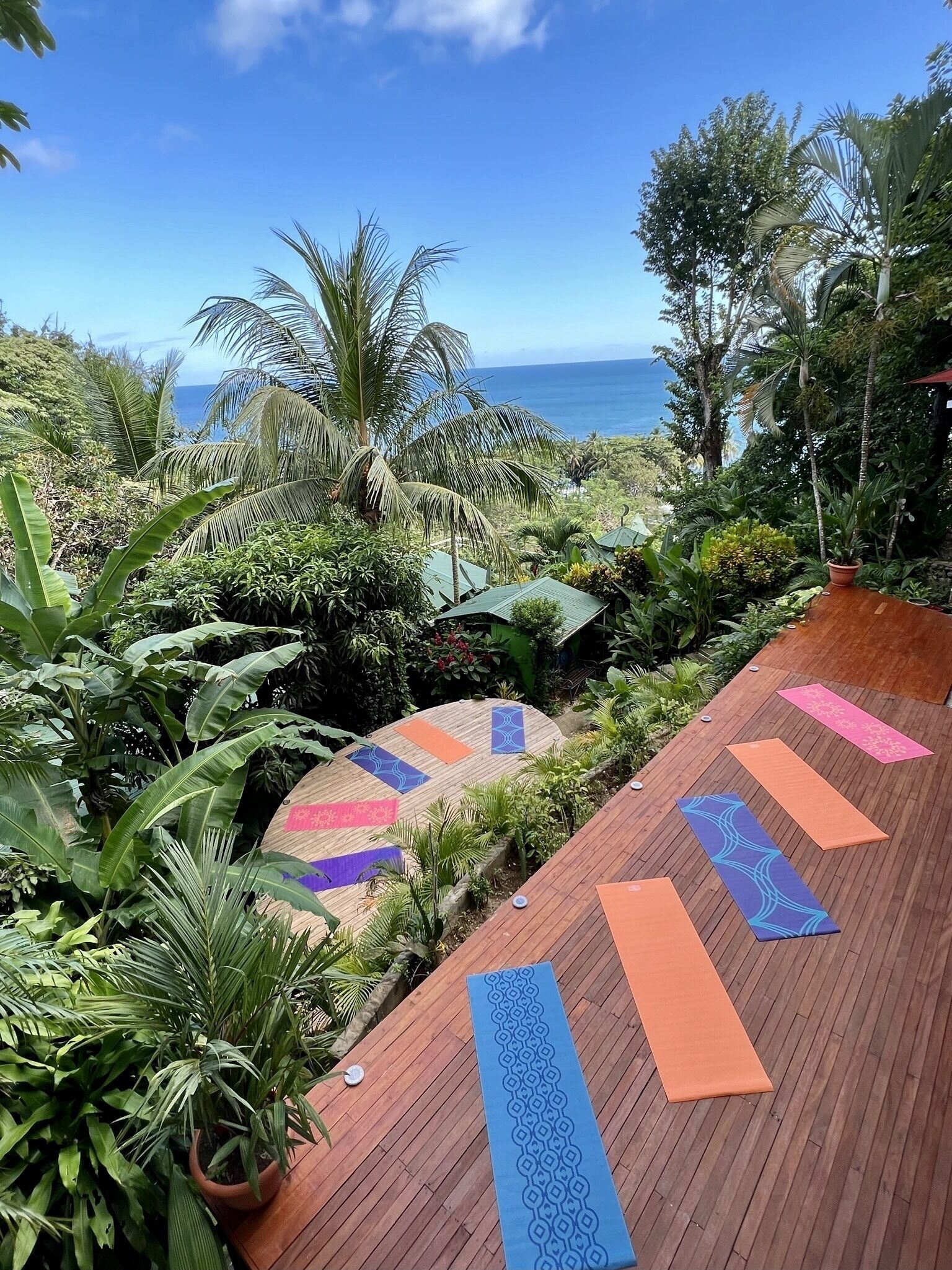 Una terraza de madera con colchonetas de yoga, rodeada de exuberante vegetación tropical y con vista a una playa.