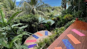 Una terraza de madera con colchonetas de yoga, rodeada de exuberante vegetación tropical y con vista a una playa.