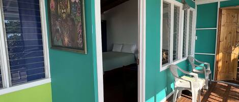 Una habitación con paredes color turquesa, suelo de madera, una cama, una ventana y una puerta que da a otra habitación.