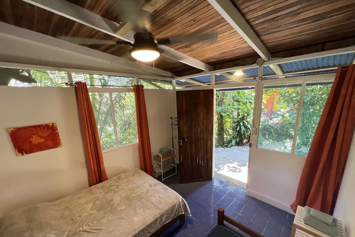 A room with a bed, a ceiling fan, a window with curtains, and a view of greenery outside.