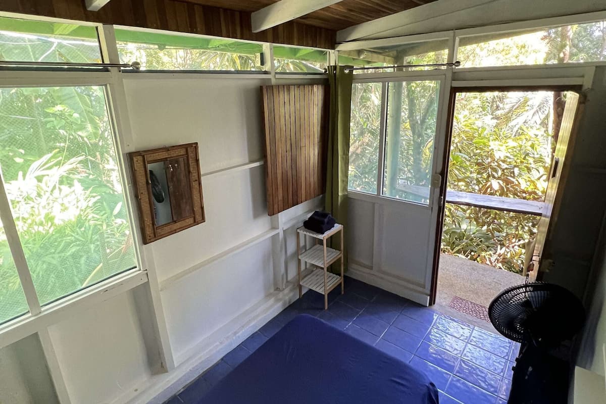 A room with a bed, a small table, a fan, and a window with a view of greenery.