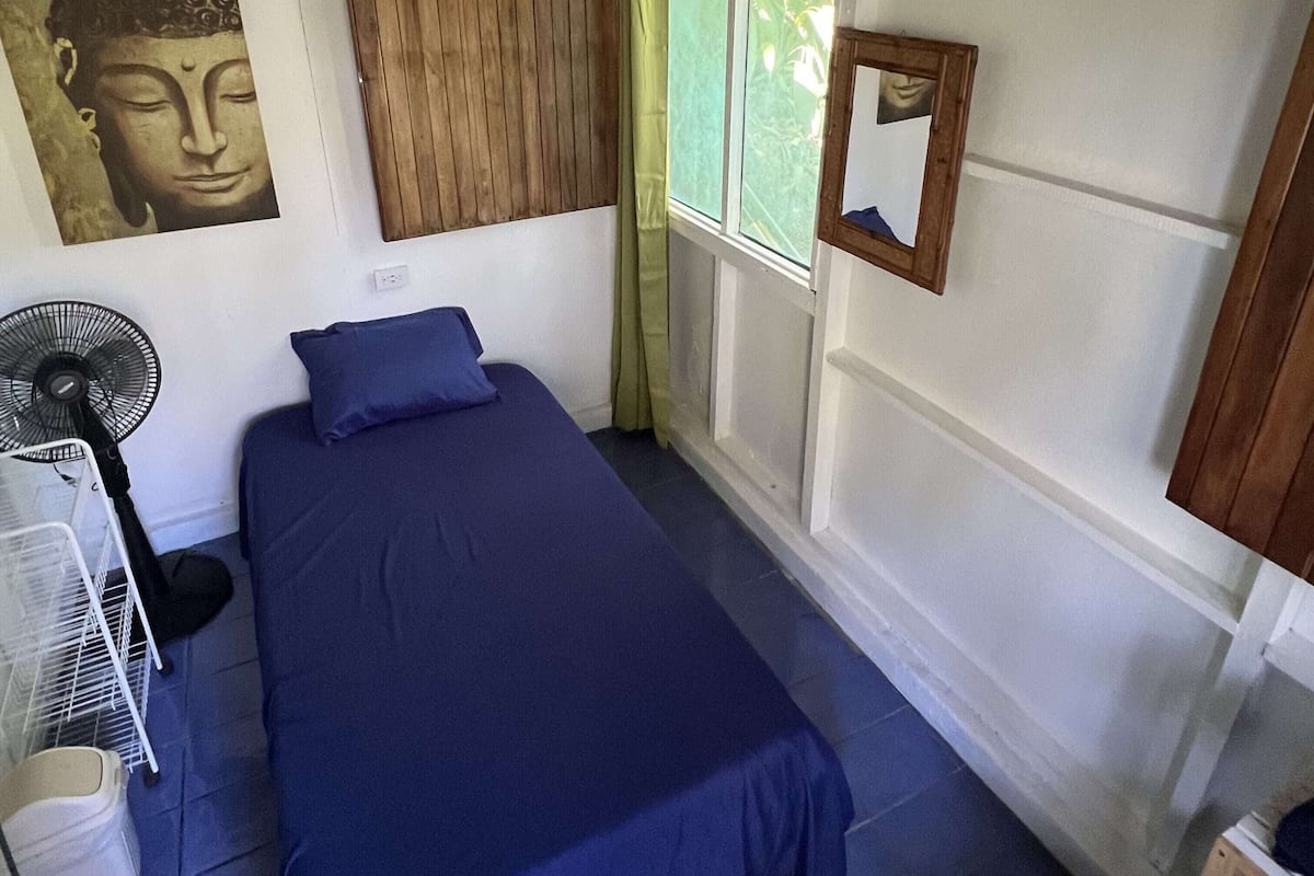A room with a bed, a fan, a window with curtains, a Buddha painting, and a small wooden chair.