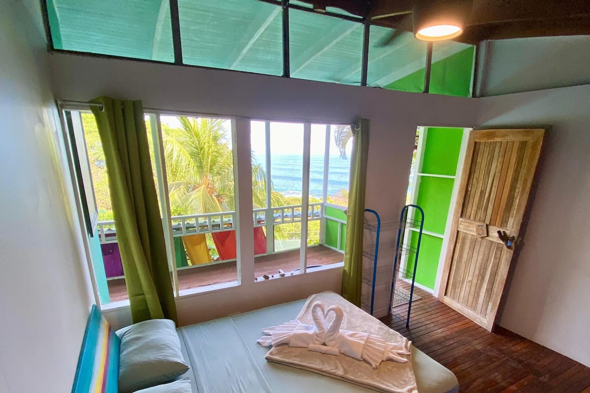 A room with a bed, a window with curtains, a view of the ocean, and a door with a number.