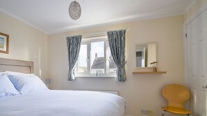 A bedroom with a bed, a chair, a window with curtains, a mirror, and a hanging light fixture. - Mead Cottage (Bridport)
