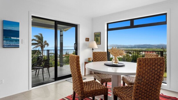 A dining area with a round table, wicker chairs, and a view of the ocean through large windows. - Kate and Harry's - Waiotahe (Opotiki)