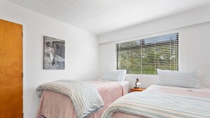 A bedroom with two beds, a wooden door, a window with blinds, and a wall-mounted artwork. - Kate and Harry's - Waiotahe (Opotiki)