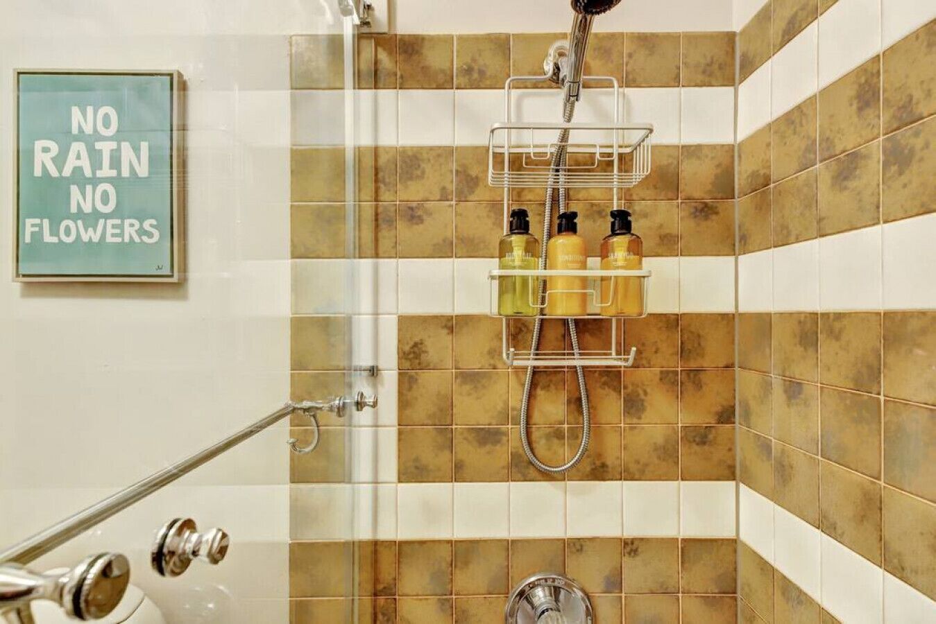 A shower area with a tiled wall, a shower caddy holding toiletries, and a 'NO RAIN NO FLOWERS' sign.