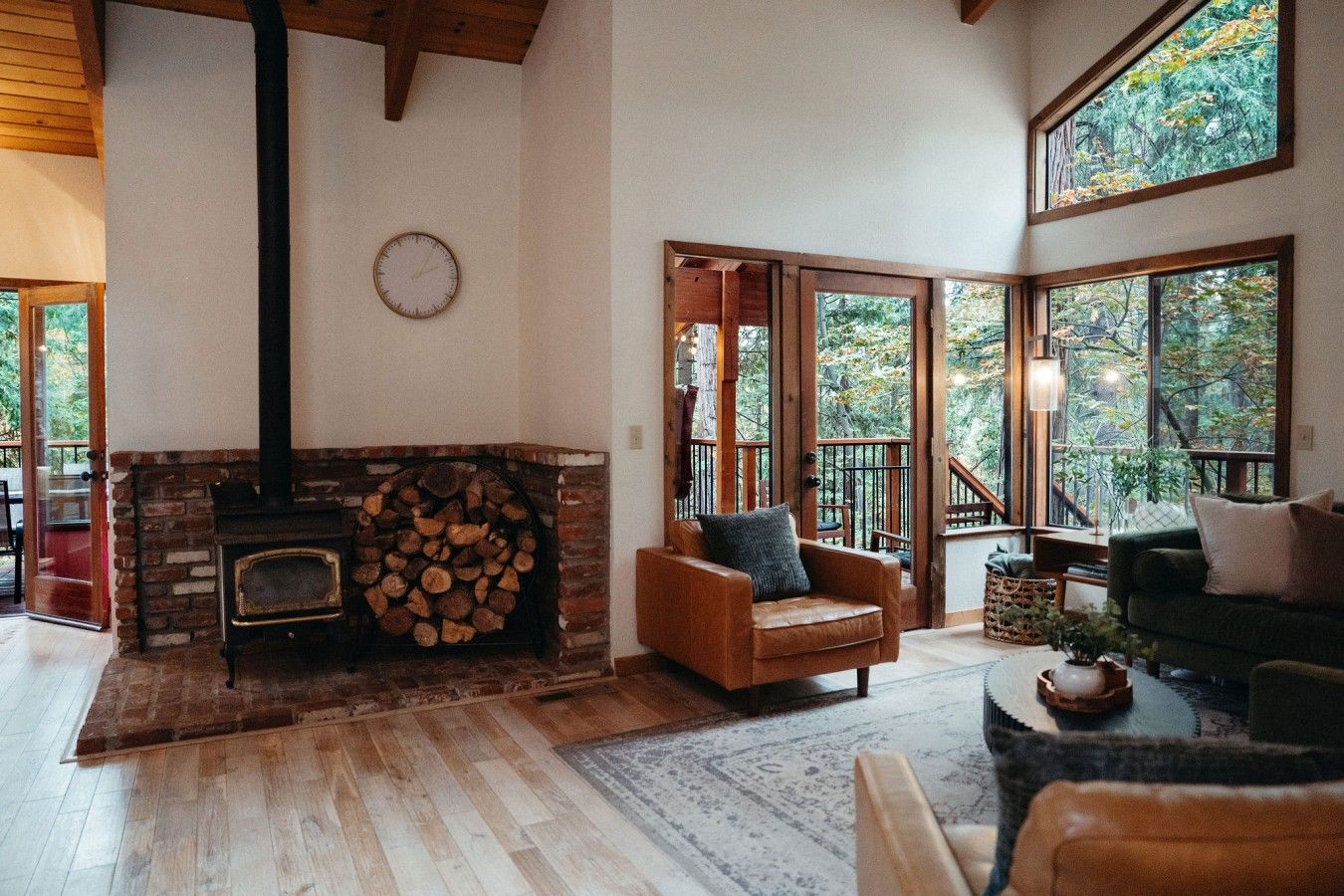Una sala de estar acogedora con chimenea, piso de madera y ventanas amplias con vistas a árboles.