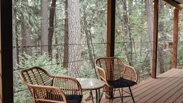 Terraza exterior cubierta con dos sillas de mimbre y una mesa, rodeada por barandilla de madera, con vista a un bosque.