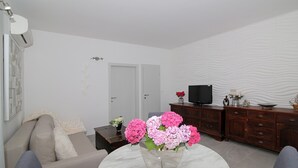 A living room with a sofa, a coffee table with a vase of flowers, a television on a wooden cabinet, and a door in the background.