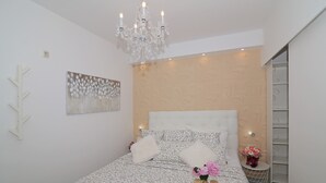 A bedroom with a large bed, a chandelier, a painting, a small table with flowers, and a white fluffy rug.