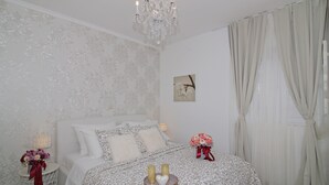 A bedroom with a bed, a chandelier, a nightstand with a lamp, and a window with curtains.