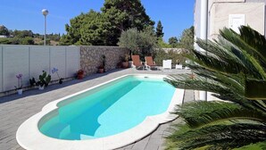 A rectangular swimming pool with a white edge, surrounded by a stone wall and potted plants.