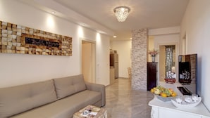A modern living room with a beige sofa, a decorative wall piece, a chandelier, and a fruit display on a white console table. - Villa Vanda - Deluxe Two Bedroom Apartment with Swimming Pool (Dubrovnik)