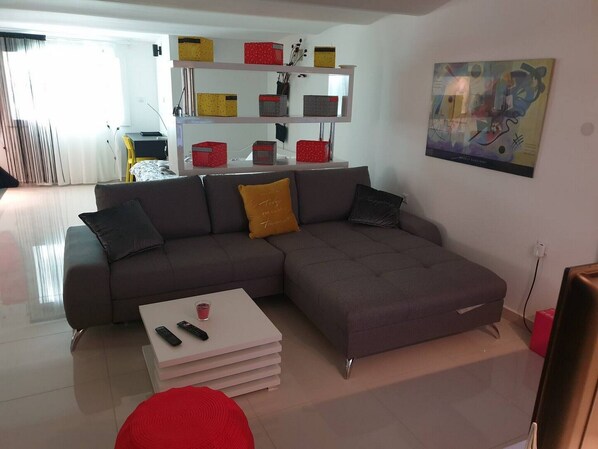 Un salon moderne avec un canapé d’angle gris, une table basse blanche avec des télécommandes, et un pouf rouge. Une étagère blanche avec des boîtes de rangement colorées se trouve à l’arrière-plan.