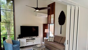 Una sala de estar con un televisor de pantalla plana, un sillón azul, un sofá marrón, una mesa de centro estampada y un ventilador de techo.