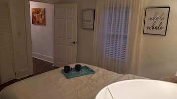 Une chambre à coucher avec un lit, une table de chevet sur laquelle reposent deux tasses, un tableau encadré accroché au mur et une fenêtre avec des rideaux.