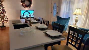 Un coin repas avec une table blanche dressée pour quatre personnes, un fauteuil bleu, un téléviseur et un sapin de Noël.
