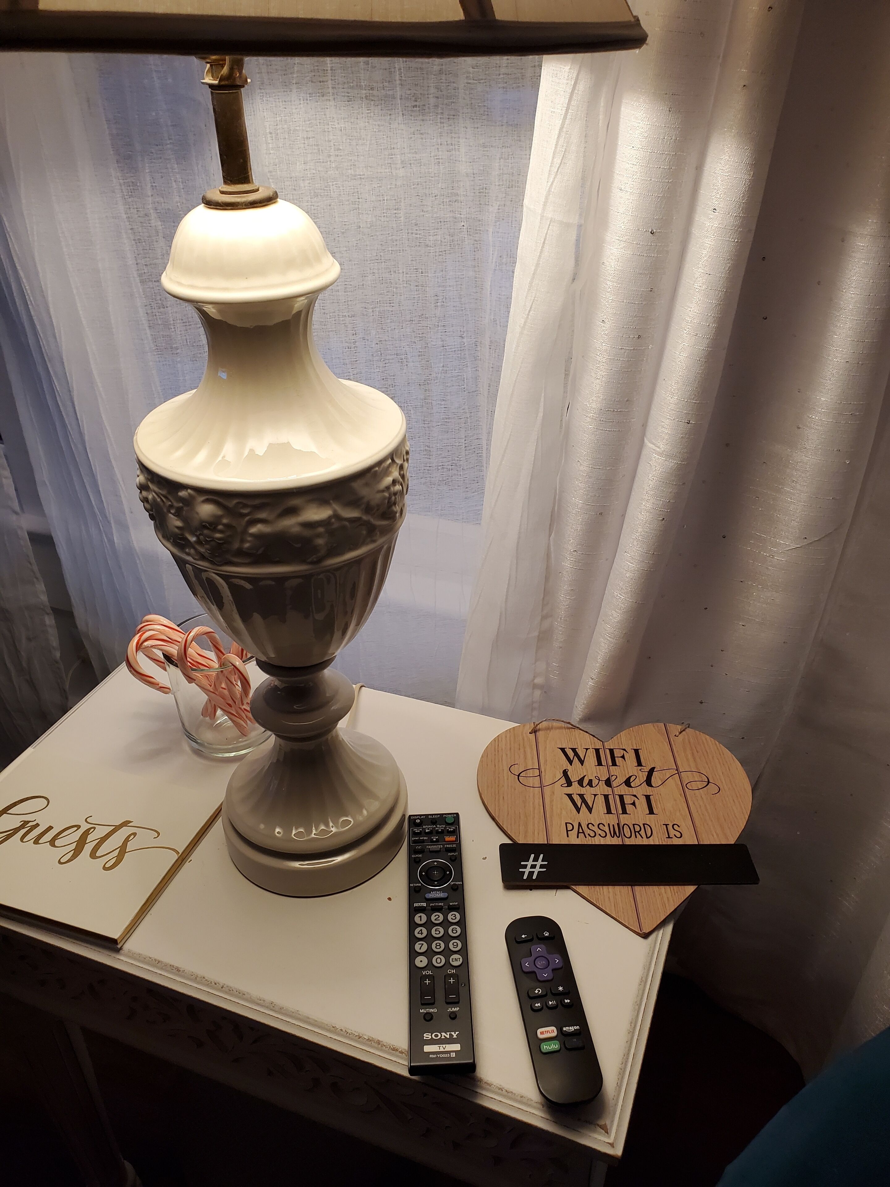 A table with a lamp, a glass container with candy canes, a Sony TV remote, a Netflix remote, and a wooden sign with a WiFi password.