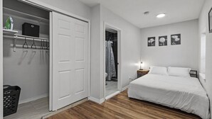 Un dormitorio con piso de madera, una cama blanca, un armario con ropa colgada y un baño con cortina de ducha.