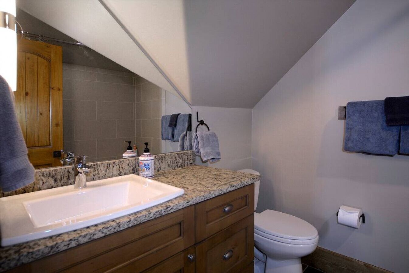 A bathroom with a granite countertop sink, wooden cabinets, a white toilet, and a towel rack with towels.