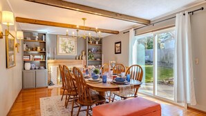 A dining area with a wooden table, chairs, and a bench. There are shelves with books and decorative items, a chandelier, and a view through a glass door.