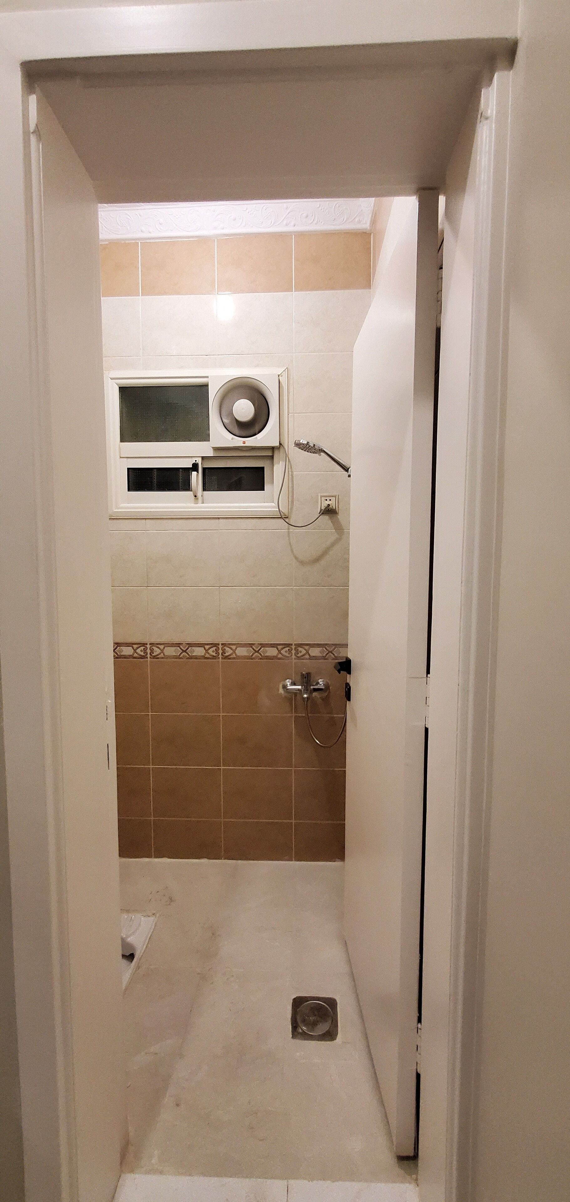 Un baño estrecho con ducha, una ventana con ventilador y paredes de azulejos.