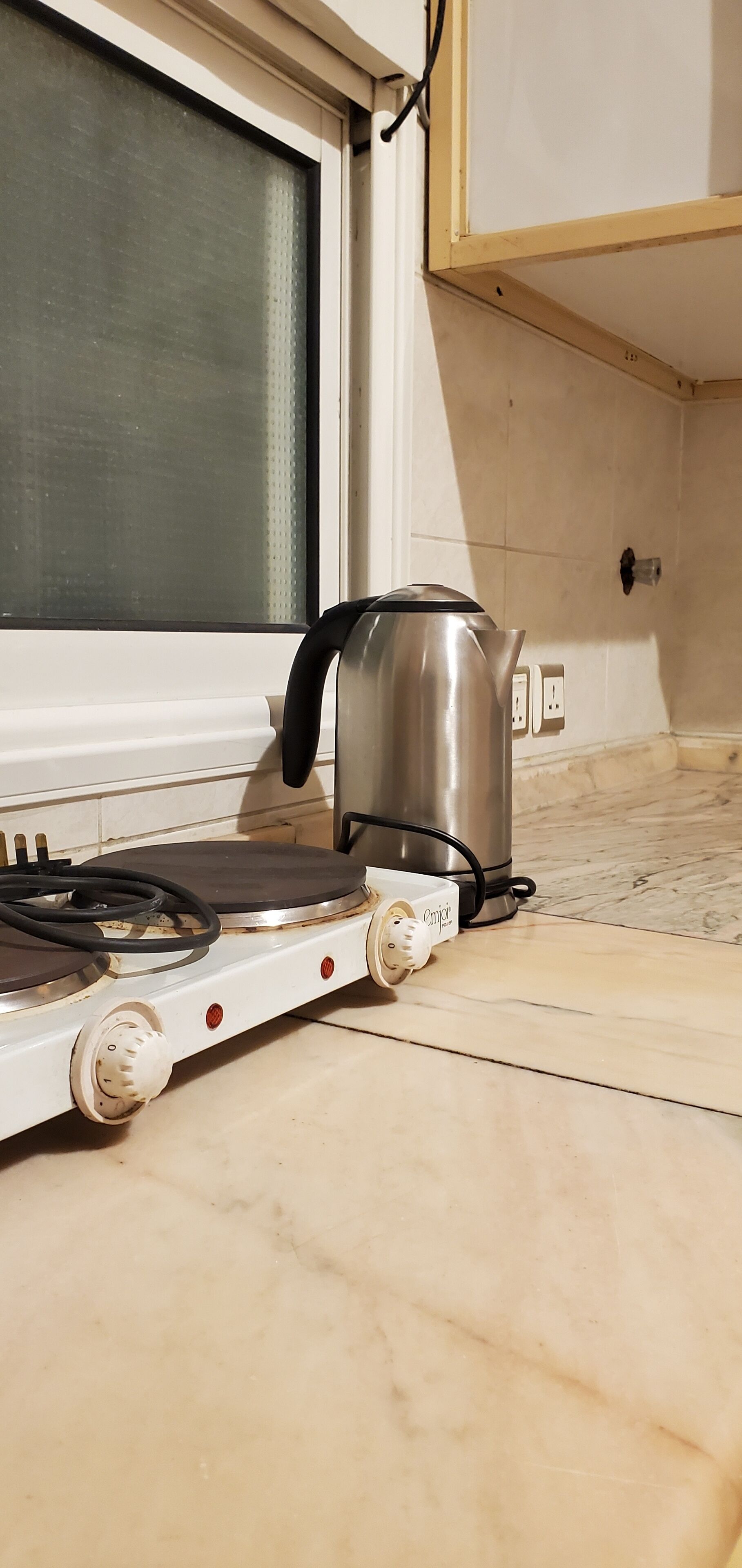 A portable electric stove with a kettle on it, placed on a tiled floor near a window.