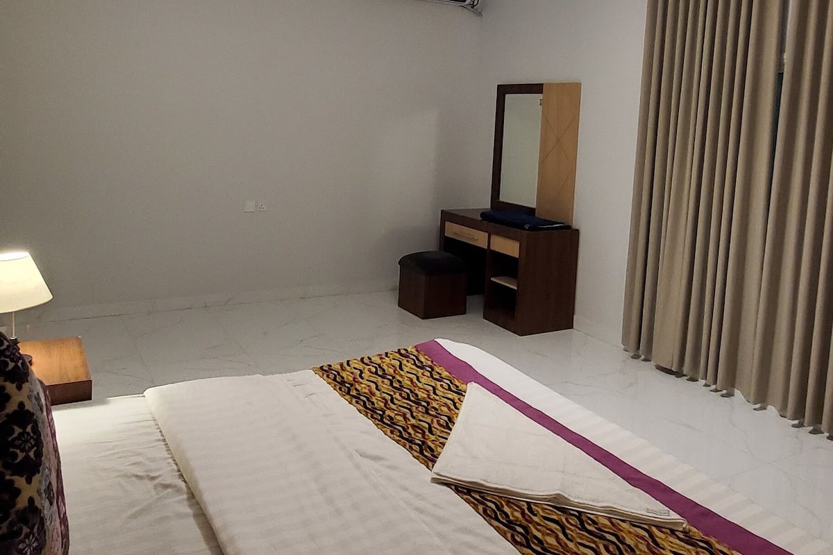 A hotel room with a bed, a bedside table with a lamp, a dresser, a mirror, and a window with curtains.