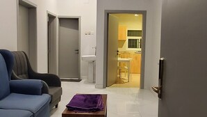 A modern living room with a blue sofa, a wooden coffee table with a purple cloth, and a kitchen visible through an open door.