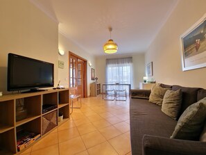 A living room with a flat-screen TV, wooden shelving, a sofa, and a dining area with a table and chairs.