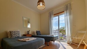 Ett sovrum med två sängar, ett stort fönster med gardiner, en hängande lampa och utsikt över omgivningarna.
