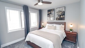Une chambre à coucher comprenant un lit, un ventilateur de plafond, deux tableaux accrochés au mur, une table de chevet avec une lampe et une fenêtre avec des stores.