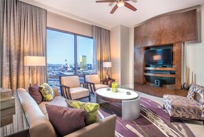 Enjoy a panoramic city view from the comfort of this stylish room, featuring a seating area with sofa and armchairs, flat-screen TV, and ceiling fan.