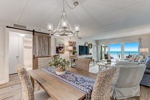 A spacious living room with a large wooden dining table, wicker chairs, a chandelier, and a view of the ocean through large windows. - Oceanfront Elegance: Your Coastal Haven (Fernandina Beach)