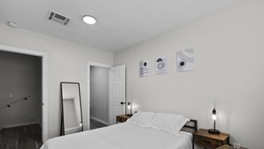 Een slaapkamer met een bed, een houten nachtkastje met een lamp, een stoel en een deur naar een andere kamer.