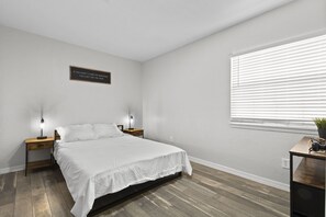 A bedroom with a bed, two bedside tables, a wall-mounted sign, a window with blinds, and a wooden floor.
