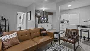 Een moderne woonkamer met een bruine leren bank, een klein rond tafeltje en een stoel met gekleurde kussens. Achtergrond is een keuken met witte kasten en een tegelwand.