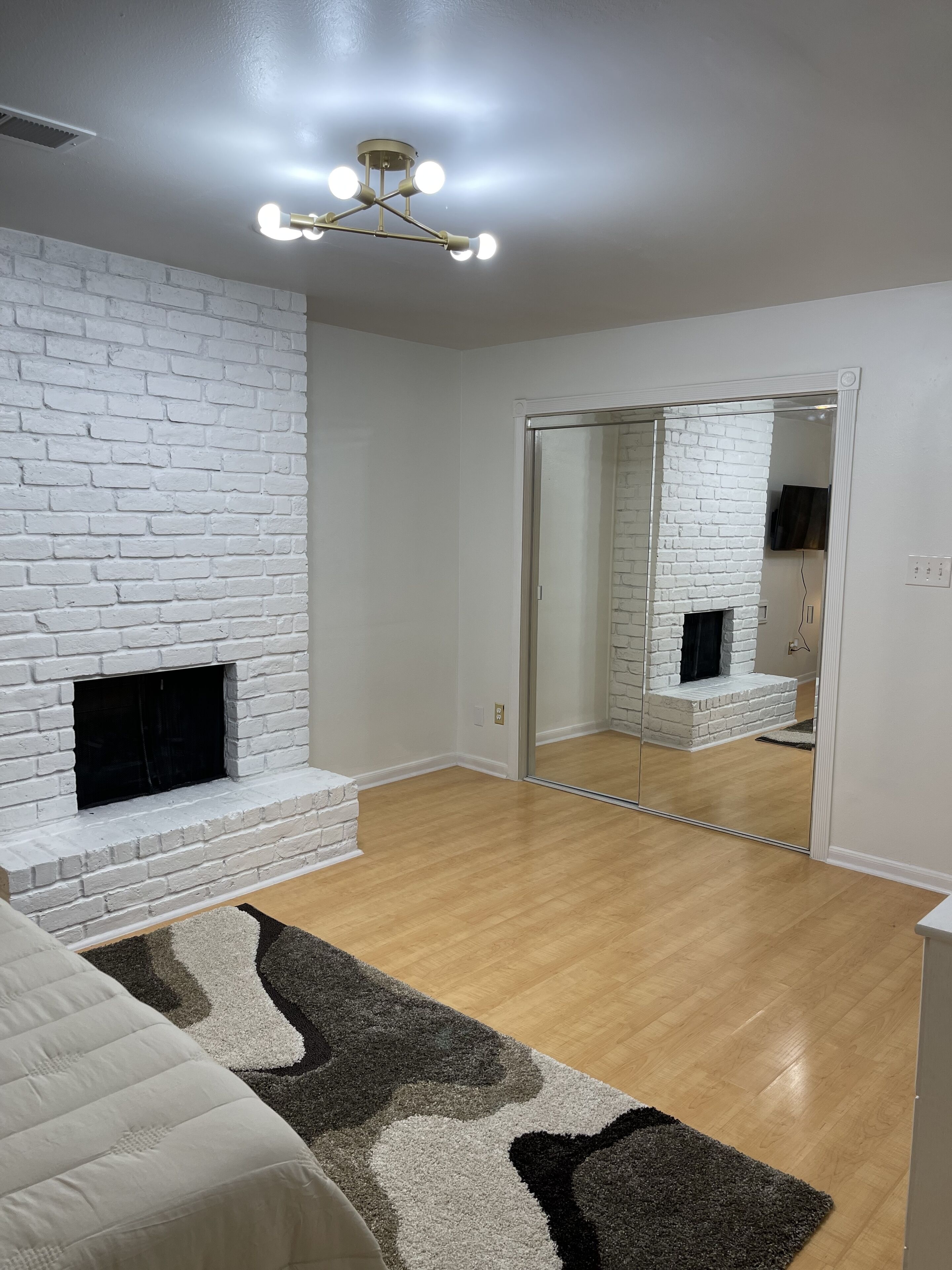 A room with a white brick fireplace, a mirrored closet door, and a light wooden floor.