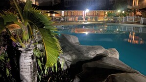 Une vue nocturne d’une piscine, avec un palmier et des rochers au premier plan, et un bâtiment doté de nombreux balcons en arrière-plan.