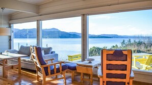 A cozy room with wooden flooring, a sofa, a chair, and a table. Large windows offer a view of a lake and mountains. - Beach front home in Nordtveitgrend with WiFi (Nordtveitgrend)