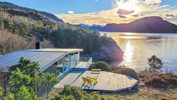 A modern house with a glass facade overlooking a lake, wooden deck with benches, and a mountainous backdrop. - Beach front home in Nordtveitgrend with WiFi (Nordtveitgrend)