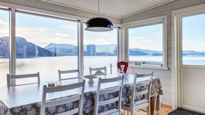 A dining area with a table and chairs facing a large window with a view of a lake and mountains. - Beach front home in Nordtveitgrend with WiFi (Nordtveitgrend)