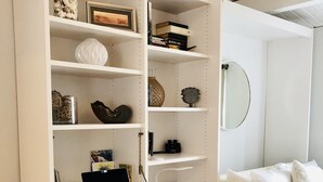 Un espacio de estudio luminoso con una computadora portátil, estanterías con libros y objetos decorativos, una silla blanca y una cama con ropa de cama blanca.