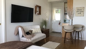 Una sala de estar acogedora con un sofá, un perro pequeño, un televisor de pantalla plana y una cocina visible a través de una puerta abierta.