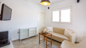 Een woonkamer met een beige bank, een klein houten tafeltje met een mand, een aan de muur bevestigde televisie, en een raam met zonwering.