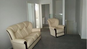 En stue med en beige sofa og lænestol, en glasdør til et badeværelse og en radiator.