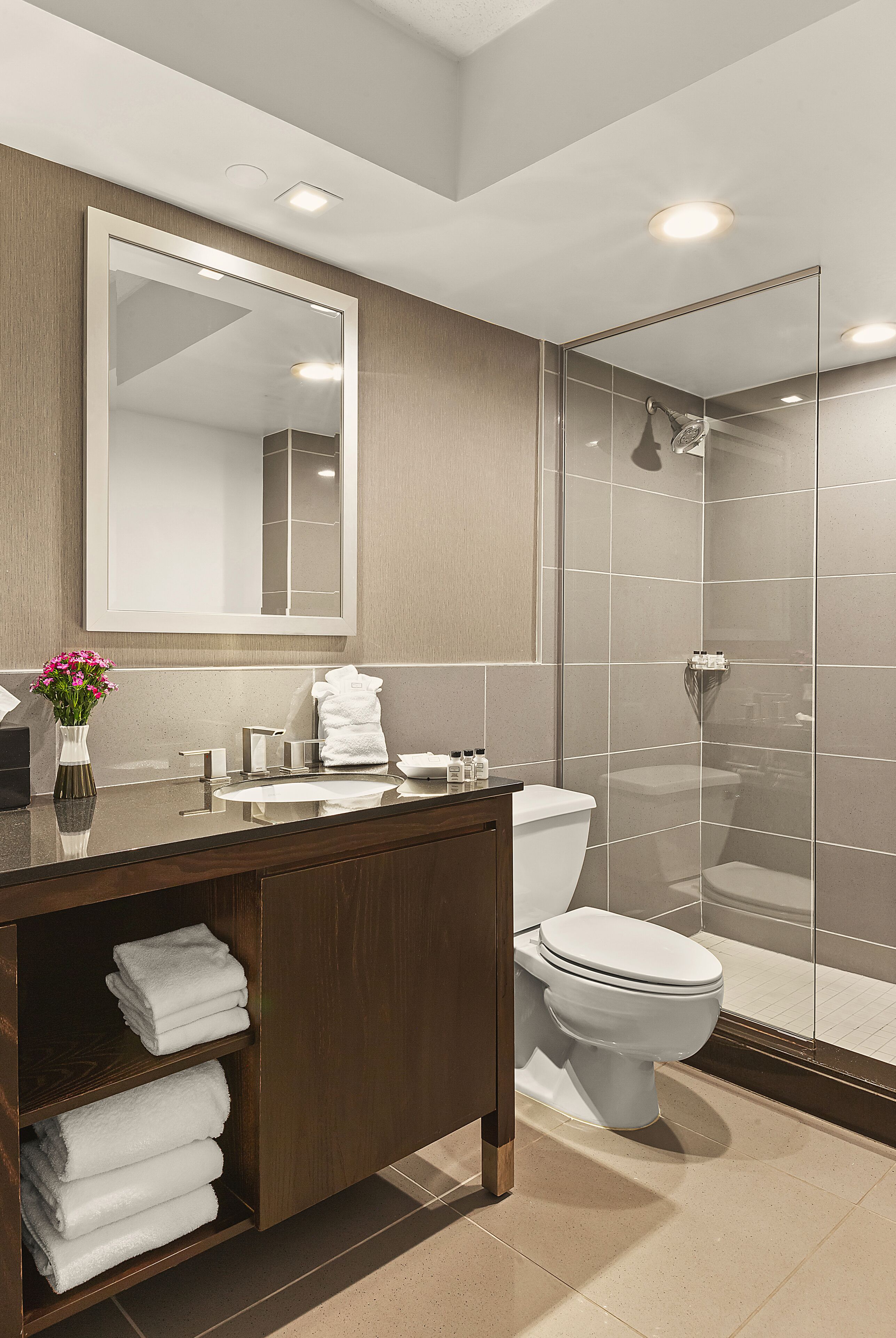 Une salle de bain moderne avec une douche à parois de verre, un grand miroir et un meuble-lavabo en bois sur lequel sont soigneusement pliées des serviettes.